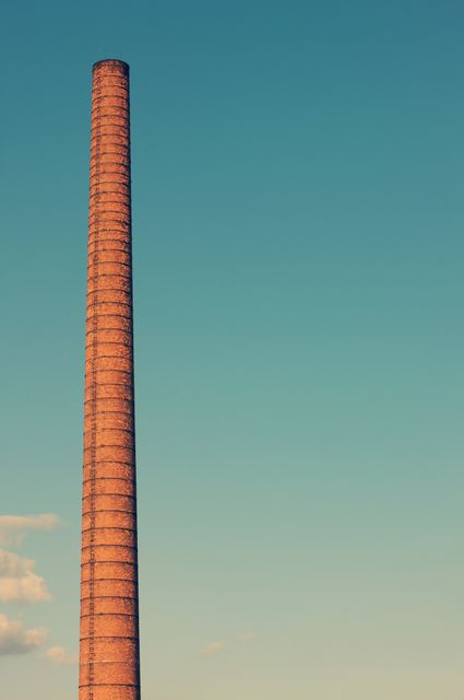 Brick chimney towering against clear blue sky captured in warm lighting. Ideal for illustrating industrial architecture, environmental topics, sustainability discussions, factory operations, or manufacturing processes.