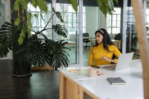 Indian woman working on laptop in modern glass-walled coworking space with plants