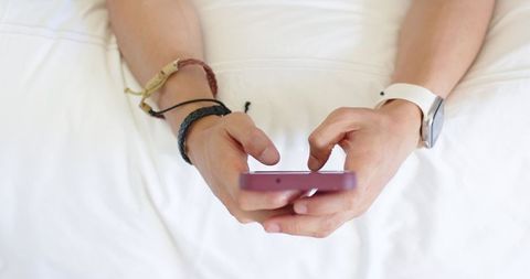 Male tapping smartphone on white bedding closeup with braided bracelets and smartwatch