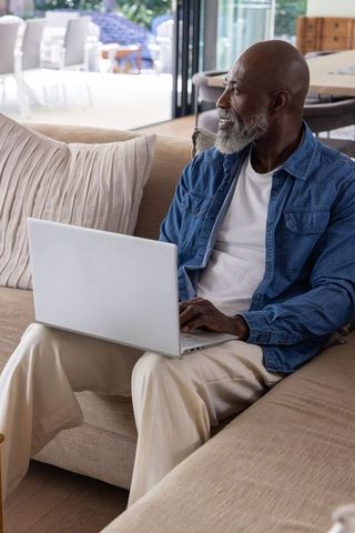 African american senior man sitting on sofa using laptop and looking toward patio at home