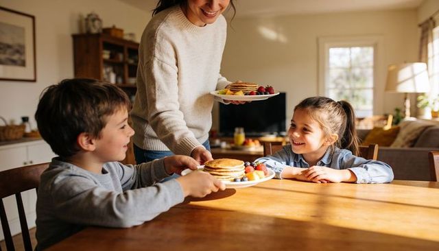 Mother serving pancakes to smiling children at sunlit kitchen table cozy family breakfast
