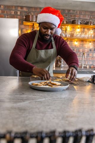 African american man wearing santa hat arranging iced cookies on cooling rack, cozy kitchen