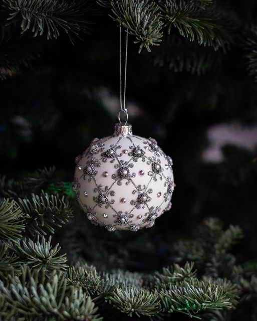 Elegant white Christmas ornament with intricate silver accents hanging on pine tree branches. Suitable for holiday greeting cards, Christmas decoration ideas, festive advertisements, or blogs about holiday preparations.