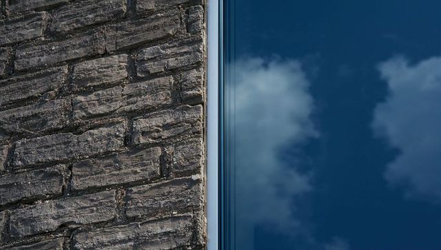 Weathered stone wall meeting reflective glass facade with sky reflection and vertical seam