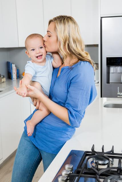 Mother holding and kissing her baby boy in a modern kitchen setting. Both are smiling, showing a moment of affection and love. Perfect for themes related to parenting, family life, modern home, and maternal bonding.