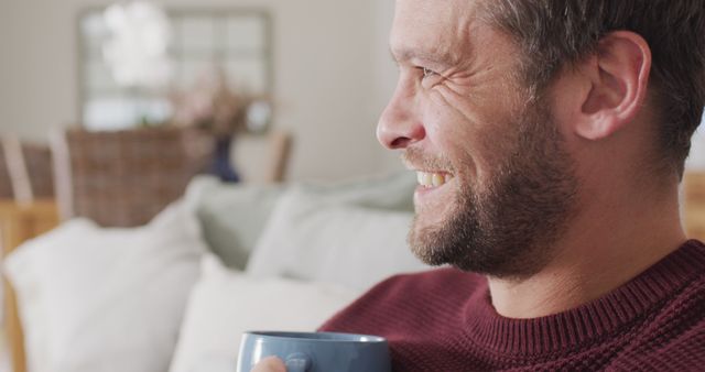 Smiling man drinking coffee, relaxing on a couch at home. Ideal for lifestyle, well-being, relaxation, comfort, and leisure concepts. Could be used in promotions related to home goods, relaxing routines, or mental well-being.