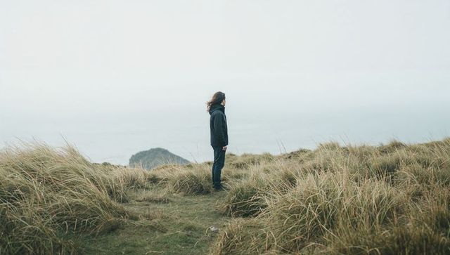 Solitary man standing on coastal dune facing foggy sea horizon, windblown grass and islet