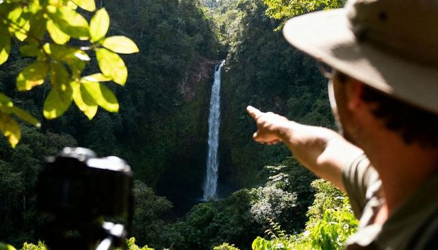 Photographer pointing toward tall tropical waterfall framed by lush rainforest canopy