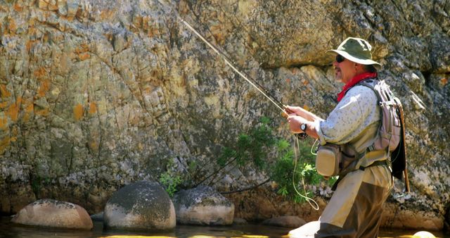 Middle-aged man fly fishing in a rocky stream situated in a mountainous landscape. He wears a wide-brim hat, a backpack, and waders. Ideal for themes related to outdoor adventures, leisure activity, camping, hiking, and fly fishing experiences.
