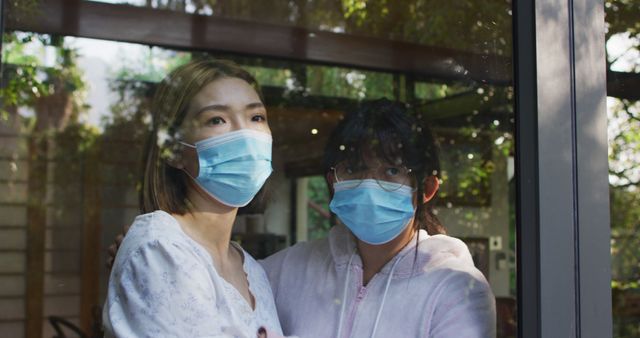 Women standing indoors with face masks, looking out through a window with reflections of trees. Suitable for concepts related to health, safety precautions, pandemic, home isolation, and personal protective measures.