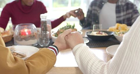 Multiracial friends holding hands around sunlit dinner table expressing togetherness