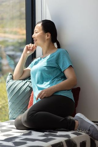 Asian woman relaxing on sunlit window seat, looking out, cozy nook, serene home lifestyle