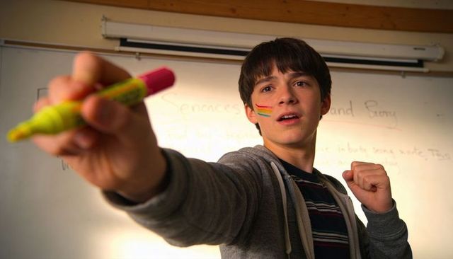 Teen presenting with highlighter and rainbow face paint at whiteboard in classroom, proud