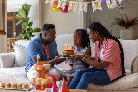 African american family celebrating daughter's birthday with rainbow cake on sofa