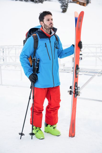 Man standing on snow covered mountain holding ski and ski poles, dressed in winter sports gear including blue jacket, red pants, and green snow boots. Ideal for use in winter sports promotions, travel brochures, outdoor adventure advertisements, and holiday travel guides.