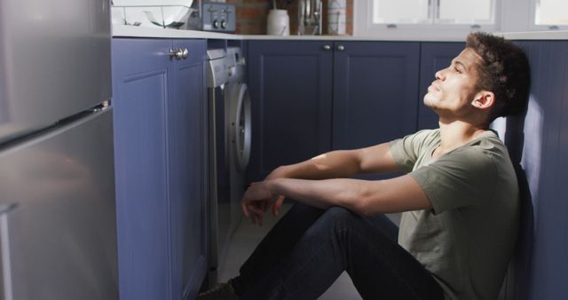 Thoughtful biracial man sitting on floor in kitchen, leaning on countertop - Download Free Stock Photos Pikwizard.com