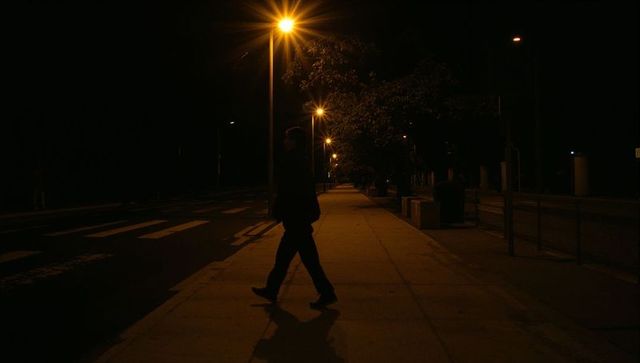 Silhouetted man walking across illuminated urban sidewalk under amber streetlights at night