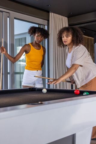 Multiracial women playing pool in modern apartment lounge, one lining up focused shot
