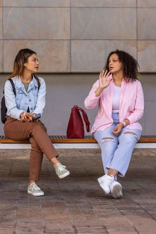 Two friends chatting on bench, african american woman gesturing, smartphone and handbag