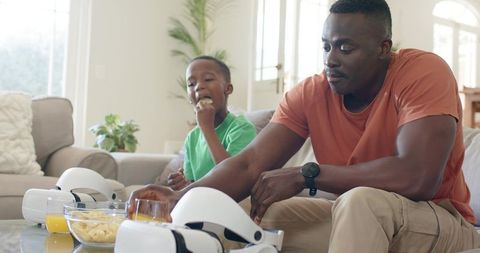 Father Reaching for VR Headset While Son Eating Chips on Couch Family Bonding at Home