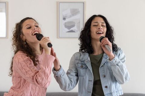 Female friends singing duet at home living room karaoke microphones cozy casual