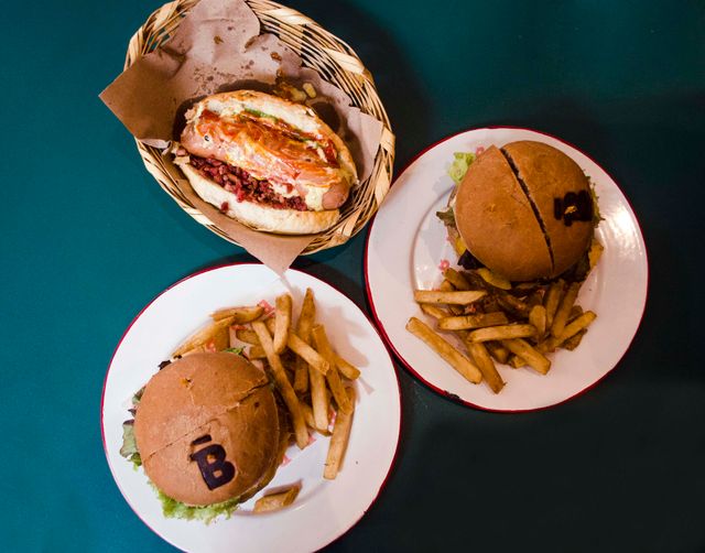 Top view of two plates with burgers and fries, and a basket with a hot dog. Placed on a green table, the burgers and hot dog appear appetizing for a meal setting photo. This image can be used for marketing restaurant menus, fast food advertisements, social media content related to food, or illustrations for articles about dining and cuisine.