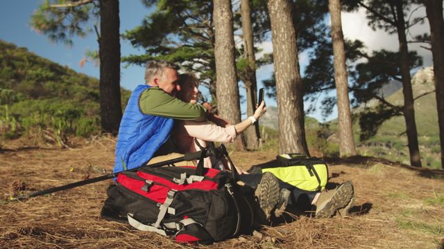 Senior couple captured enjoying a hike in nature, sitting on a grassy meadow with backpacks and Nordic walking sticks. They are seen taking a selfie, smiling and enjoying each other's company. Ideal for use in promotional material for outdoor activities, senior fitness programs, travel agencies, and retirement lifestyle advertisements.