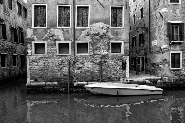 An old, abandoned boat is docked on a calm canal with aged brick buildings in the background. This black and white scene captures the timeless and serene essence of an urban European setting. Ideal for use in travel brochures, historical presentations, architectural studies, or vintage-themed projects.