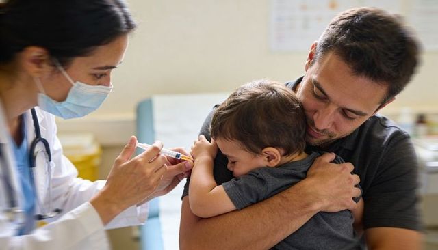 Masked clinician vaccinating tearful toddler while father comforting during pediatric clinic visit