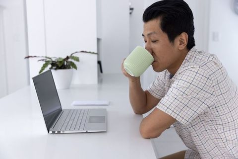 Sipping pale green mug while working on laptop at minimalist white kitchen counter