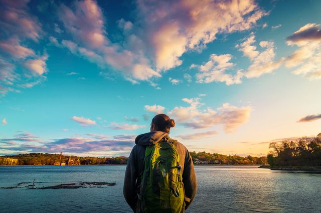 A person with a green backpack stands by a calm lake, admiring a colorful sunset with vibrant clouds in the sky. This image can be used for travel blogs, outdoor activity advertisements, nature appreciation websites, and motivational content encouraging outdoor adventure and exploration.