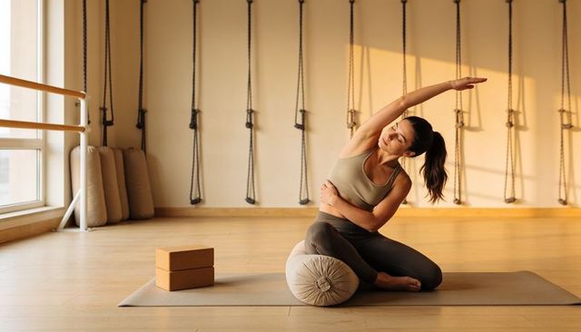 Woman practicing side stretch on bolster in sunlit yoga studio with cork blocks
