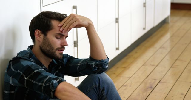 Young man wearing casual plaid shirt sitting on wooden floor in kitchen with hand on forehead, appearing stressed. Useful for articles discussing mental health, stress management, emotional well-being, and men's mental health. Suitable for use in blogs, social media posts, and healthcare content.