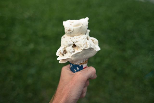 Hand holding ice cream cone with patriotic wrapping outdoors. Ideal for themes related to summer, American holidays, desserts, and outdoor activities. Perfect for marketing materials, social media posts, and advertisements focusing on festive celebrations and delicious treats.