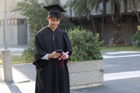 Asian male graduate checking phone on campus wearing cap and gown holding diploma