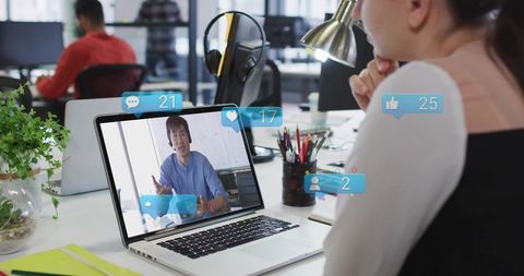 Woman watching virtual meeting on laptop with live social reactions in open-plan office