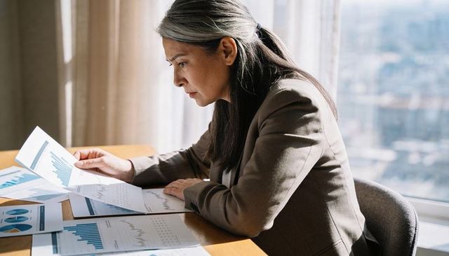 Middle-aged asian businesswoman reviewing financial charts and reports at sunlit office desk