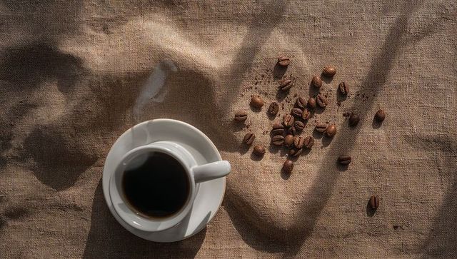 Steaming cup of black coffee on burlap with scattered roasted coffee beans