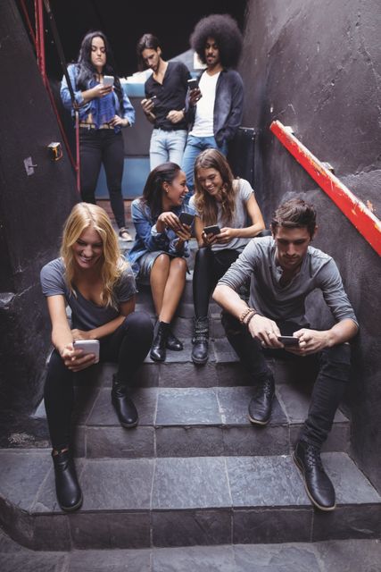 Group of young friends sitting on steps in a nightclub, all engaged with their mobile phones. Ideal for illustrating themes of social media, technology, modern communication, nightlife, and youth culture. Perfect for use in articles, advertisements, and social media posts related to technology, social interactions, and urban lifestyle.