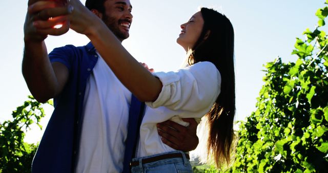 A diverse couple is enjoying a romantic moment outdoors, sharing a toast with glasses of wine, with copy space. Their joyful expressions and the vineyard setting suggest a celebration or a leisurely date in a picturesque location.