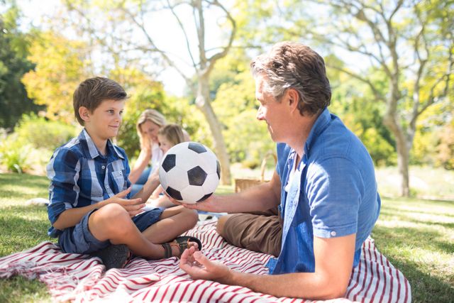 Father and son enjoying quality time together in a park, playing with a football on a sunny day. Ideal for use in family-oriented advertisements, parenting blogs, outdoor activity promotions, and lifestyle articles focusing on family bonding and leisure activities.