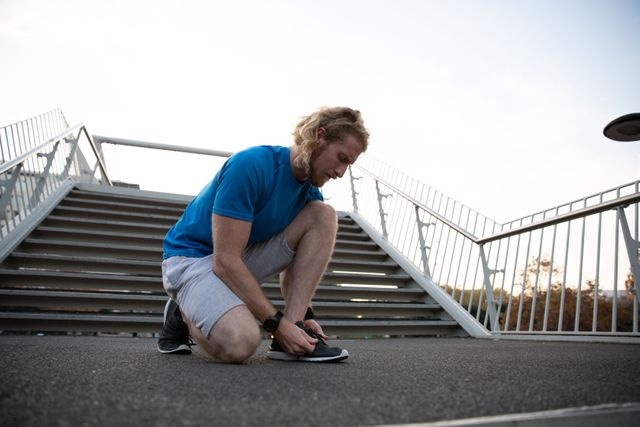 Low angle front view of a fit Caucasian man with long blonde hair wearing sportswear exercising outdoors in the city on a sunny day with blue sky, kneeling and tying his shoe.