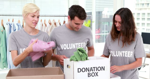 Two women and a man, all young adults and Caucasian, are sorting clothes into a donation box, with copy space. They are volunteers engaged in a charitable activity, contributing to a clothing drive.