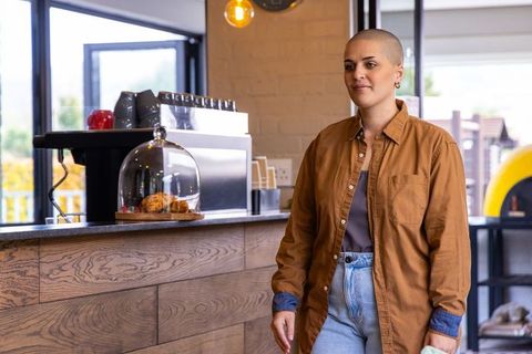 Confident woman walking through sunlit cafe near espresso machine and pastry display