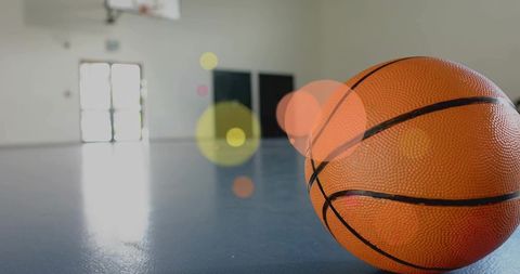 Basketball resting on shiny indoor court, hoop visible, atmospheric bokeh lighting