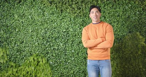 Confident asian man standing with arms crossed against lush green ivy wall casual lifestyle
