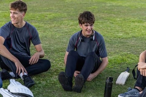 Young cricketers sitting on outfield laughing during practice with gear and headphones