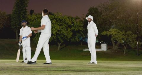 Floodlit night cricket scene with players standing on pitch and indian batsman holding bat