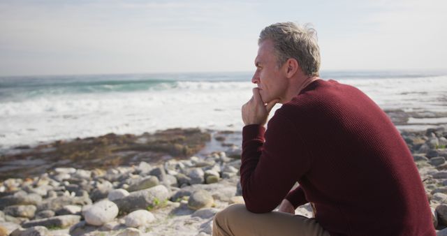 Elderly man sitting on rocky shore looking out at the ocean, appearing deep in thought. Use for themes of meditation, reflection, nature's beauty, loneliness, or retirement lifestyle.