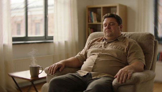 Middle-aged man relaxing in armchair with steaming mug by window, cozy calm home moment